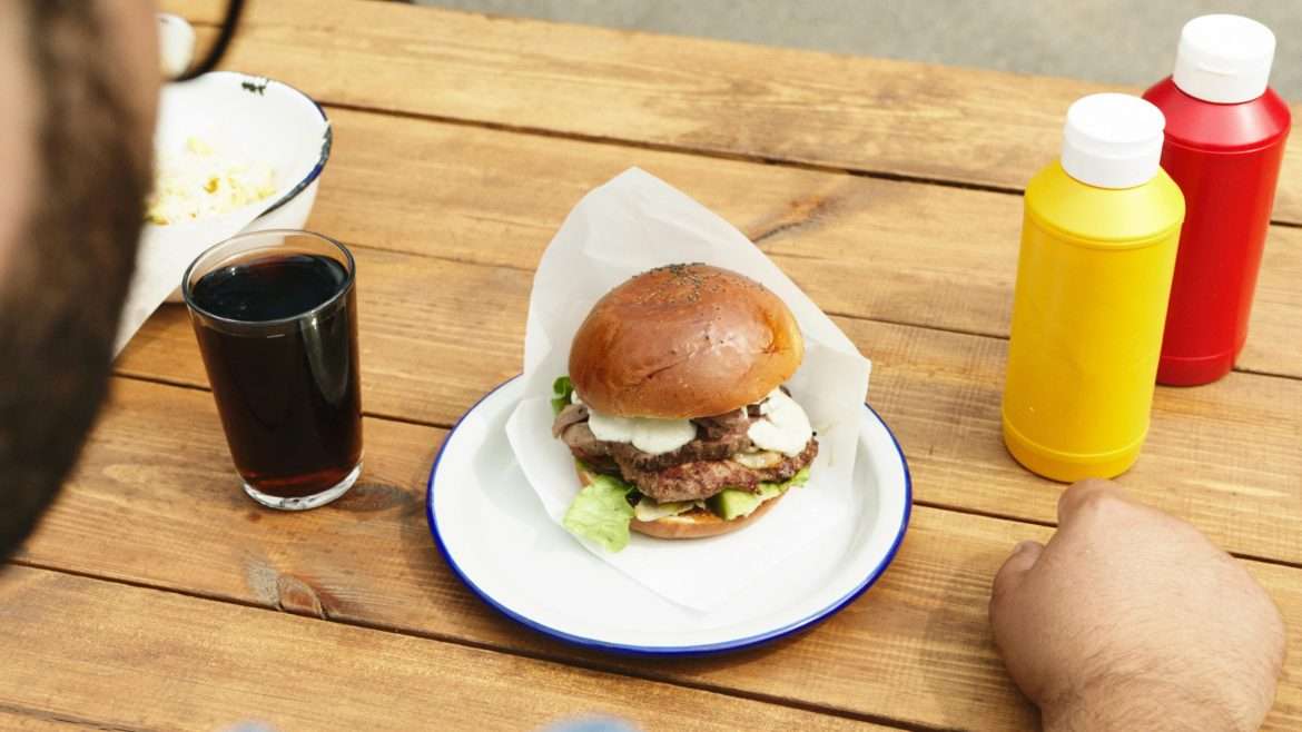 Close-up of a cheeseburger with lettuce, accompanied by soda and condiments on a wooden table.